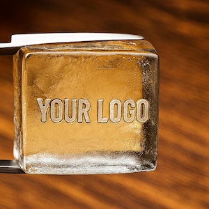 May include: A clear ice cube with the text "YOUR LOGO" engraved on its surface, held by metal tongs. The ice cube has a golden hue and sits against a blurred wooden background.