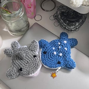 May include: Two handmade crochet coin purses. One is gray with black accents, the other is blue with white dots. Both have a zipper closure and a star-shaped charm. The purses are shaped like a cartoon animal.