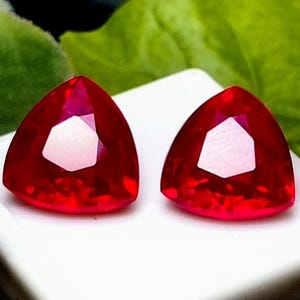 May include: Two red triangular gemstones with a faceted cut. The gemstones are set on a white surface.