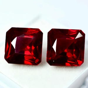 Puede incluir: Dos gemas rojo oscuro, talladas en forma cuadrada. Las gemas tienen un diseño facetado, que captura la luz y muestra brillantes reflejos internos. Las piedras están sobre un fondo blanco, resaltando su rico color y claridad.