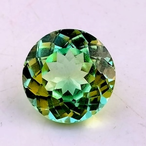 May include: A round, faceted green gemstone with a star-shaped pattern in the center. The gemstone has a yellow hue around the edges.