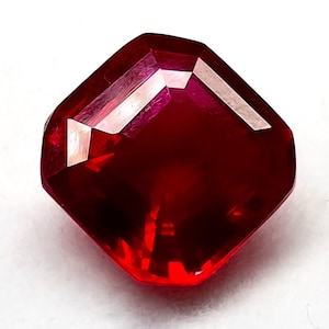 Puede incluir: Una piedra preciosa de color rojo intenso con forma cuadrada y muchas facetas. La piedra preciosa está tallada en forma de cojín.