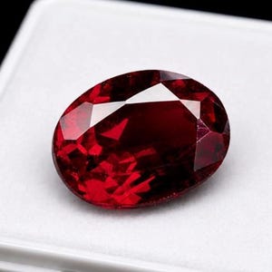 May include: A deep red, oval-cut gemstone with multiple facets that reflect light. The stone displays a rich, saturated crimson hue and is positioned on a white surface, showcasing its transparent quality and precise geometric cutting style.