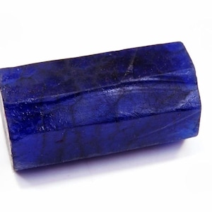 May include: A rectangular, deep blue gemstone with a faceted surface. The stone has a polished appearance and a rich, saturated colour. The gemstone is set against a white background, highlighting its colour and texture. The object appears to be a raw, uncut stone.