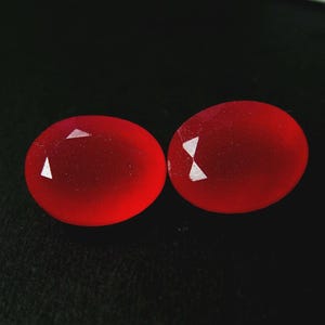 Puede incluir: Dos gemas rojas, ovaladas y facetadas, se muestran sobre un fondo negro. Las gemas tienen un aspecto pulido y translúcido, con algunas facetas reflectantes que captan la luz. Las piedras son de tamaño y forma similares.