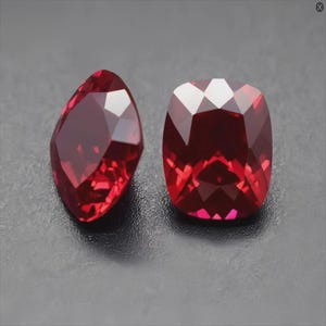 Könnte beinhalten: Zwei rote Kissen-Schliff-Edelsteine, einer etwas größer als der andere, werden auf einer dunkelgrauen Oberfläche präsentiert. Die Edelsteine sind facettiert und haben einen hohen Glanz.