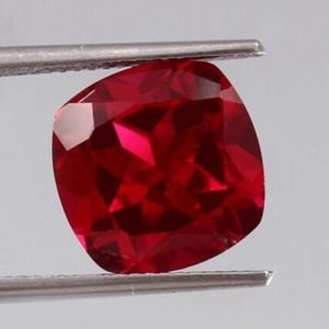 May include: A deep red gemstone with a cushion cut. The gemstone is held in a pair of tweezers.