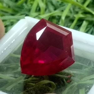 Puede incluir: Una piedra preciosa de color rojo intenso con un corte facetado en forma de corazón. La piedra preciosa se muestra en una caja blanca con un fondo verde.