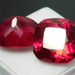 Könnte beinhalten: Zwei facettierte, kissenförmige, rote Edelsteine. Die Edelsteine sind ein tiefes, sattes Rot mit brillanten Facetten, die das Licht reflektieren. Die Edelsteine werden auf einer weißen Oberfläche vor einem dunklen Hintergrund präsentiert.