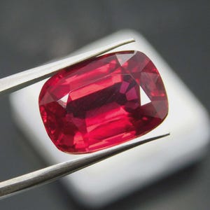 Pode incluir: Uma pedra preciosa vermelha escura, facetada e de formato retangular, é segurada por pinças prateadas. A gema possui múltiplas facetas que refletem a luz, criando uma exibição brilhante de cor. O fundo é branco e cinza neutro.