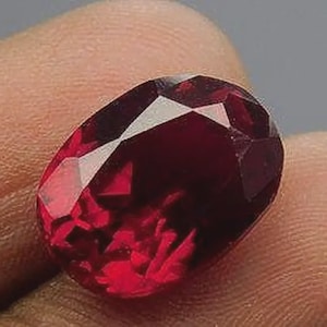 Pode incluir: Uma gema oval de cor vermelha escura com corte facetado. A gema está engastada em um anel e está sendo segurada por uma mão.