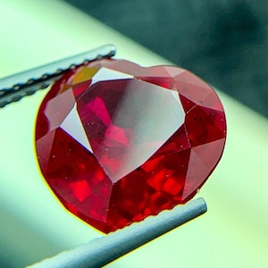 Könnte beinhalten: Ein herzförmiger, facettierter, tiefroter Edelstein wird von einer Pinzette gehalten. Der Edelstein hat einen Brillantschliff, der das Licht reflektiert und seine satte Farbe zur Geltung bringt. Der Hintergrund ist ein weiches, verschwommenes Grün.