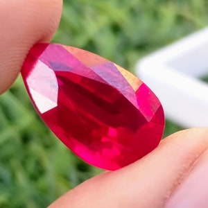 May include: A deep red pear-shaped gemstone with a faceted cut. The gemstone is held in a hand, with a green background.