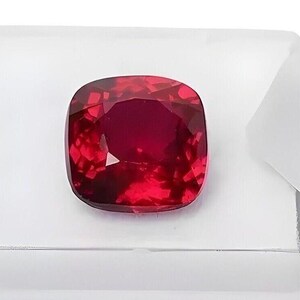 Könnte beinhalten: Ein tiefroter Edelstein mit einem Kissen-Schliff. Der Edelstein ist auf einem weißen Hintergrund platziert.
