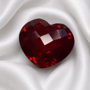 May include: A deep red, heart-shaped gemstone featuring a faceted surface that reflects light. The stone has a polished, multi-faceted cut with sharp edges and a glossy finish, displaying varying shades of crimson and dark red throughout its structure.