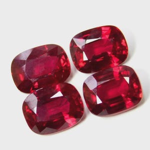 Könnte beinhalten: Vier kissenförmig geschliffene Rubine mit tiefroter Farbe werden vor einem weißen Hintergrund präsentiert. Die Edelsteine haben Facetten, die das Licht reflektieren und einen funkelnden Effekt erzeugen. Die Anordnung zeigt die Klarheit und Farbe der Edelsteine.