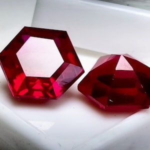 Könnte beinhalten: Zwei rote Edelsteine, einer hexagonal und einer achteckig, mit einem facettierten Schliff. Die Edelsteine ​​werden auf einer weißen Oberfläche präsentiert.