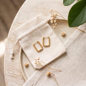 May include: A pair of gold-toned rectangular hoop earrings with a smooth finish, resting on a small, off-white fabric drawstring pouch. The earrings feature a hinged closure mechanism and are positioned centrally on the textured cloth surface.