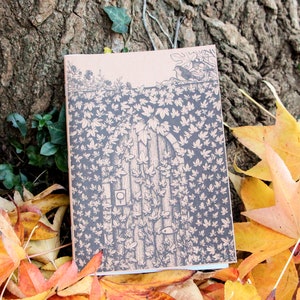 May include: A small, brown notebook with a black and white illustration of a door covered in leaves. The notebook is lying on a bed of fallen autumn leaves.