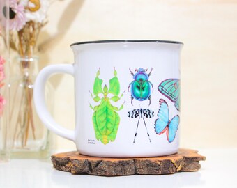 Puede incluir: Taza de cerámica blanca con borde negro que presenta una ilustración colorida de insectos, incluyendo un insecto hoja verde, un escarabajo verde, una libélula y dos mariposas. El texto "Melissa Graves" está impreso en la taza.