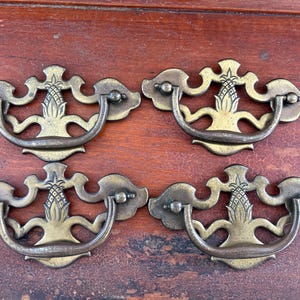 May include: Four antique brass drawer pulls with a pineapple design in the center. The pulls have a curved handle and decorative backplates. The brass has a warm, aged patina, suggesting vintage or antique furniture hardware.