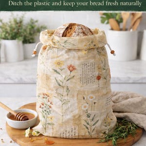 May include: A reusable beeswax bread bag filled with a loaf of bread. The bag is beige with a floral pattern and a drawstring closure. Text on the bag reads "Reusable Beeswax Bread Bag" and "Ditch the plastic and keep your bread fresh naturally."