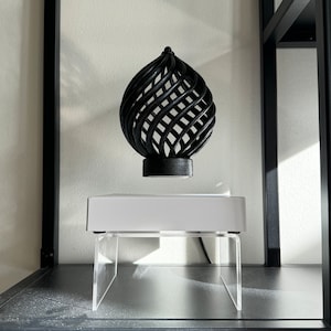 Puede incluir: Una escultura de orbe geométrico de madera negra que levita sobre una base blanca con patas de acrílico transparente. La escultura está suspendida en el aire en un estante.