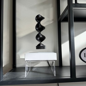 Könnte beinhalten: Eine schwarze Holzskulptur in Spiralform schwebt über einer weißen Plattform mit transparenten Acrylbeinen. Die Skulptur befindet sich auf einem schwarzen Regal.