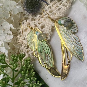 May include: A pair of gold-toned earrings with a delicate, wing-like design. The wings feature a gradient of green and blue hues, accented with small, light-colored gemstones. The earrings are set against a backdrop of white flowers and lace.