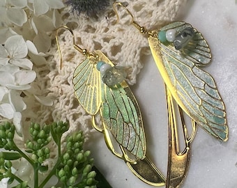 Puede incluir: Un par de pendientes dorados con un diseño delicado en forma de ala. Las alas presentan un degradado de tonos verdes y azules, acentuado con pequeñas piedras preciosas de color claro. Los pendientes se colocan sobre un fondo de flores blancas y encaje.