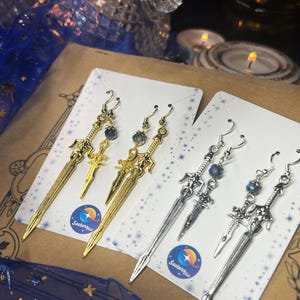 Könnte beinhalten: Ohrringe mit Schwertdesign in Gold- und Silbertönen. Jedes Paar hat einen kleinen Schwertanhänger und eine blaue Perle. Die Ohrringe werden auf weißen Karten mit einem Logo präsentiert. Die Ohrringe sind ca. 10 cm lang.