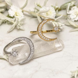 Peut inclure: Deux bagues ouvertes avec un motif de croissant de lune et d'étoile. Une bague est dorée et l'autre est argentée. Les deux bagues sont incrustées de petites pierres claires le long de la lune et présentent une étoile avec une pierre centrale.