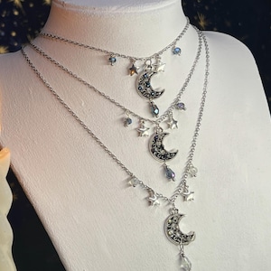 Könnte beinhalten: Dreilagige silberne Halsketten mit Mond- und Sternanhängern. Jede Halskette hat eine andere Länge und ein anderes Design mit baumelnden Kristallen und Perlen. Die Halsketten werden auf einer weißen Mannequin-Büste ausgestellt.