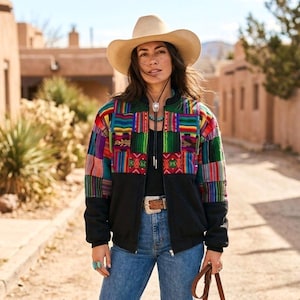 May include: A beige cowboy hat, a black bomber jacket with multicolored woven fabric panels on the upper chest and sleeves, a black shirt, blue jeans, a brown leather belt with a silver buckle, and a bolo tie with a turquoise stone accent.