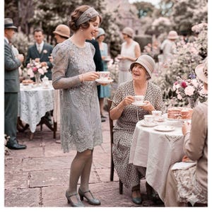 Könnte beinhalten: Eine Vintage-Teeparty-Szene im Freien. Eine Frau in einem grauen Spitzenkleid und Stirnband hält eine Teetasse und lächelt. Eine andere Frau in einem gemusterten Kleid sitzt an einem Tisch mit Kuchen und Blumen. Weitere Gäste sind im Hintergrund zu sehen.