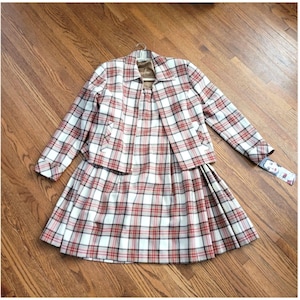 May include: A vintage plaid skirt suit in red, white, and black. The set includes a collared jacket and a pleated skirt. The jacket has long sleeves and pockets. The skirt falls to the knee. A tag is attached to the sleeve.