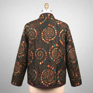 May include: A dark brown quilted jacket featuring a repeating pattern of spiral designs composed of small, circular patches in shades of orange, yellow, teal, and brown. The jacket has long sleeves, a high collar, and a dark purple trim along the edges.