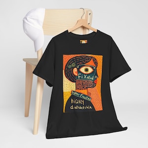 May include: Black t-shirt featuring a colourful graphic of a stylized face. The face has an eye, and the words "Fixated", "hyper fixated", and "highly distractible" are printed on the shirt. A white baseball cap rests on a wooden chair.