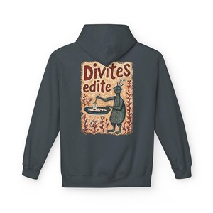 May include: Dark grey hoodie with a whimsical illustration on the back. The artwork features a cartoon creature holding a spoon over a dish, with the words "Divites edite" above. The design is set against a backdrop of stylized plants.