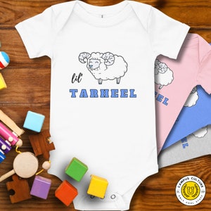 Onesie (white) with lil Tarheel text and fluffy ram with blue heart nose.  Alternate colors layered behind (pink, blue, grey). Laying on wood background with plush moon and star accent.