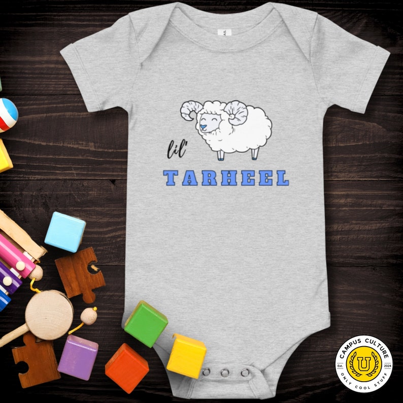 Short sleeve one piece Lil Tarheel text on grey onesie with fluffy ram with blue heart nose  | Team Mascot | Newborn | School Spirit. On wood background with bright scattered toys.