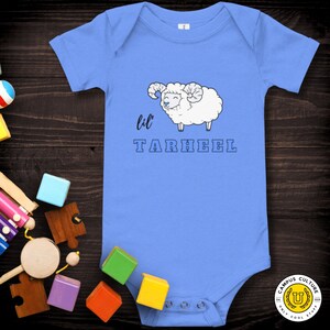 Short sleeve one piece Lil Tarheel text on blue onesie with fluffy ram with blue heart nose  | Team Mascot | Newborn | School Spirit. On wood background with bright scattered toys.