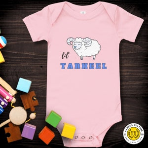 Short sleeve one piece Lil Tarheel text on pink onesie with fluffy ram with blue heart nose  | Team Mascot | Newborn | School Spirit. On wood background with bright scattered toys.