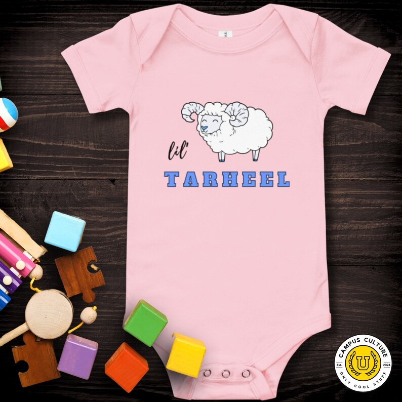 Short sleeve one piece Lil Tarheel text on pink onesie with fluffy ram with blue heart nose  | Team Mascot | Newborn | School Spirit. On wood background with bright scattered toys.
