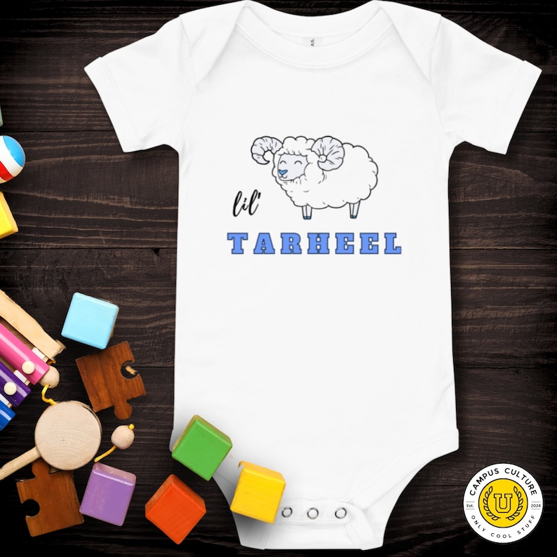 Short sleeve one piece Lil Tarheel text on white onesie with fluffy ram with blue heart nose  | Team Mascot | Newborn | School Spirit. On wood background with bright scattered toys. UNC