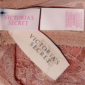 May include: Two pink and white Victoria's Secret tags with the text "Victoria's Secret" on them. The tags are on a pink and white lace fabric.