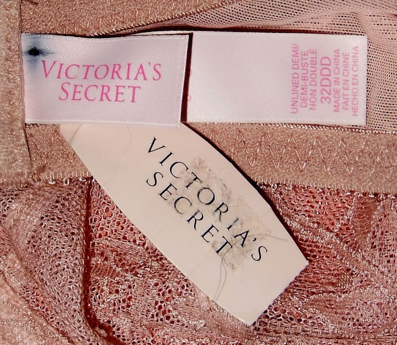May include: Two pink and white Victoria's Secret tags with the text "Victoria's Secret" on them. The tags are on a pink and white lace fabric.