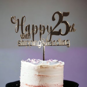 Op de afbeelding: Gouden taarttopper met de tekst "Happy 25th with shipping & handling" op een witte taart met roze glazuur.