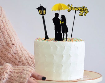 Può includere: Un topper per torta con la sagoma nera di una coppia sotto un ombrello giallo accanto a un lampione nero. Il topper include il testo Yasmin 27 in caratteri gialli e neri, posizionato su una torta bianca glassata con codette colorate.