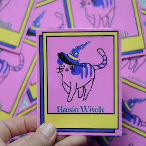 May include: A pink card with a yellow border and blue trim features a cartoon cat wearing a purple and blue witch hat. The cat has blue stripes and is standing on a pink background. The text "Basic Witch" is printed in blue on a yellow background.